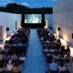 Schermo all'aperto in piazza gremita di spettatori, locandine di film e luci serali.